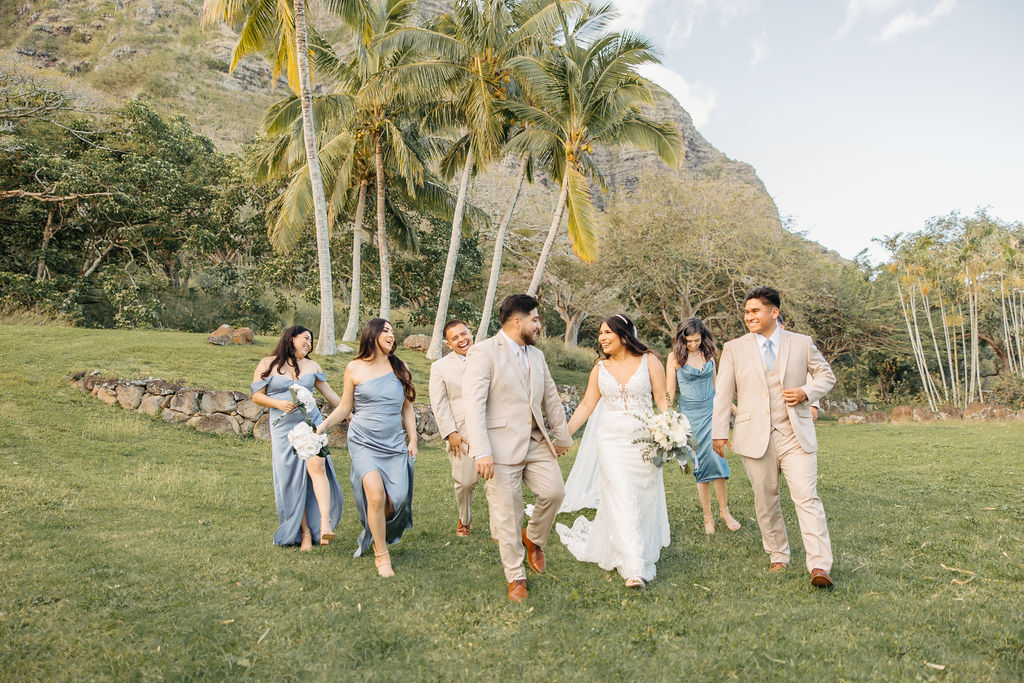 A couple in wedding attire kisses outdoors, surrounded by a cheering wedding party dressed in blue and beige. Palm trees and a hill are in the background.