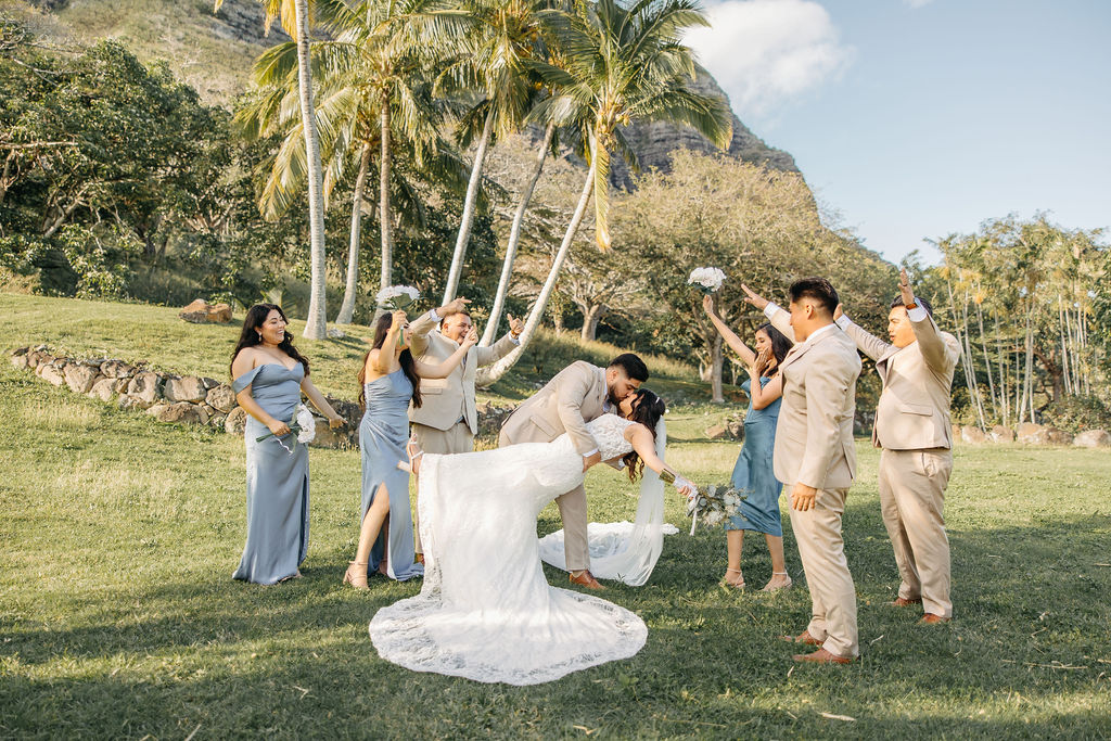 A couple in wedding attire kisses outdoors, surrounded by a cheering wedding party dressed in blue and beige. Palm trees and a hill are in the background.