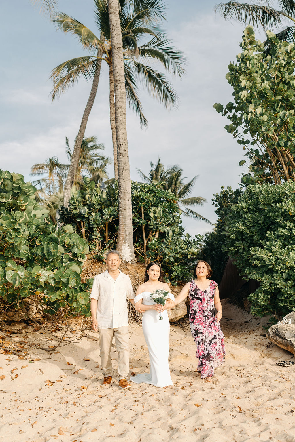 A couple stands holding hands during a beach wedding ceremony, with an officiant and a guitarist in the background by the ocean.