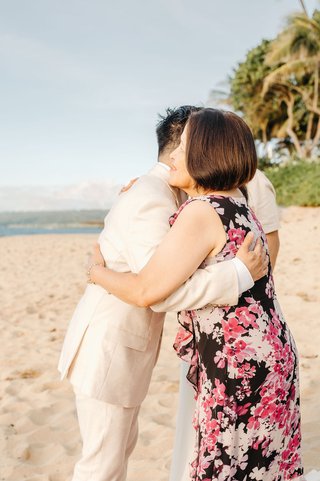 A couple stands holding hands during a beach wedding ceremony, with an officiant and a guitarist in the background by the ocean.