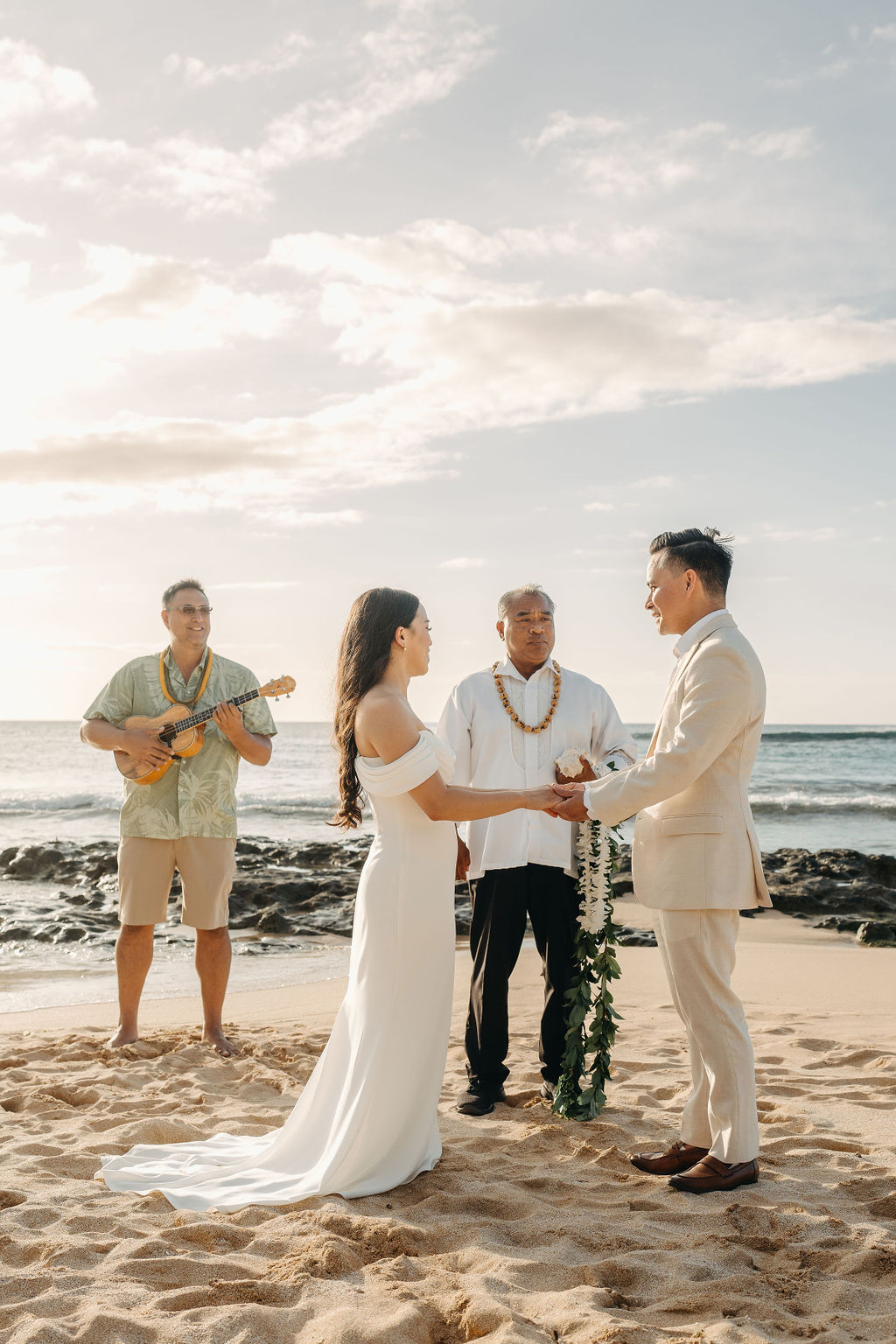 A couple stands holding hands during a beach wedding ceremony, with an officiant and a guitarist in the background by the ocean.