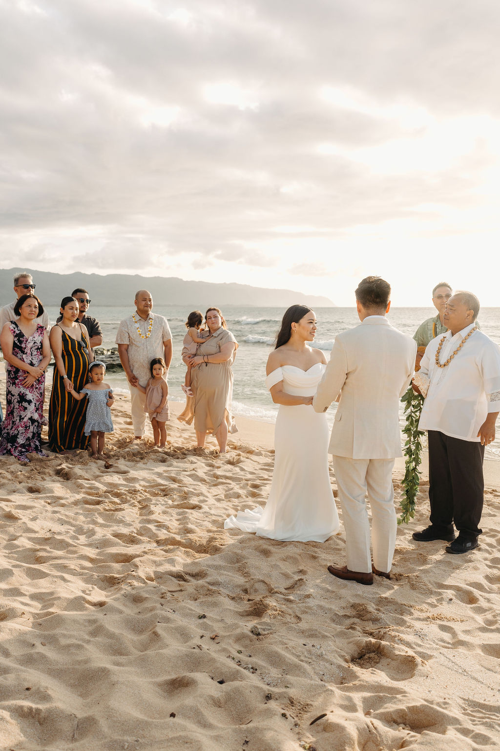 A couple stands holding hands during a beach wedding ceremony, with an officiant and a guitarist in the background by the ocean.