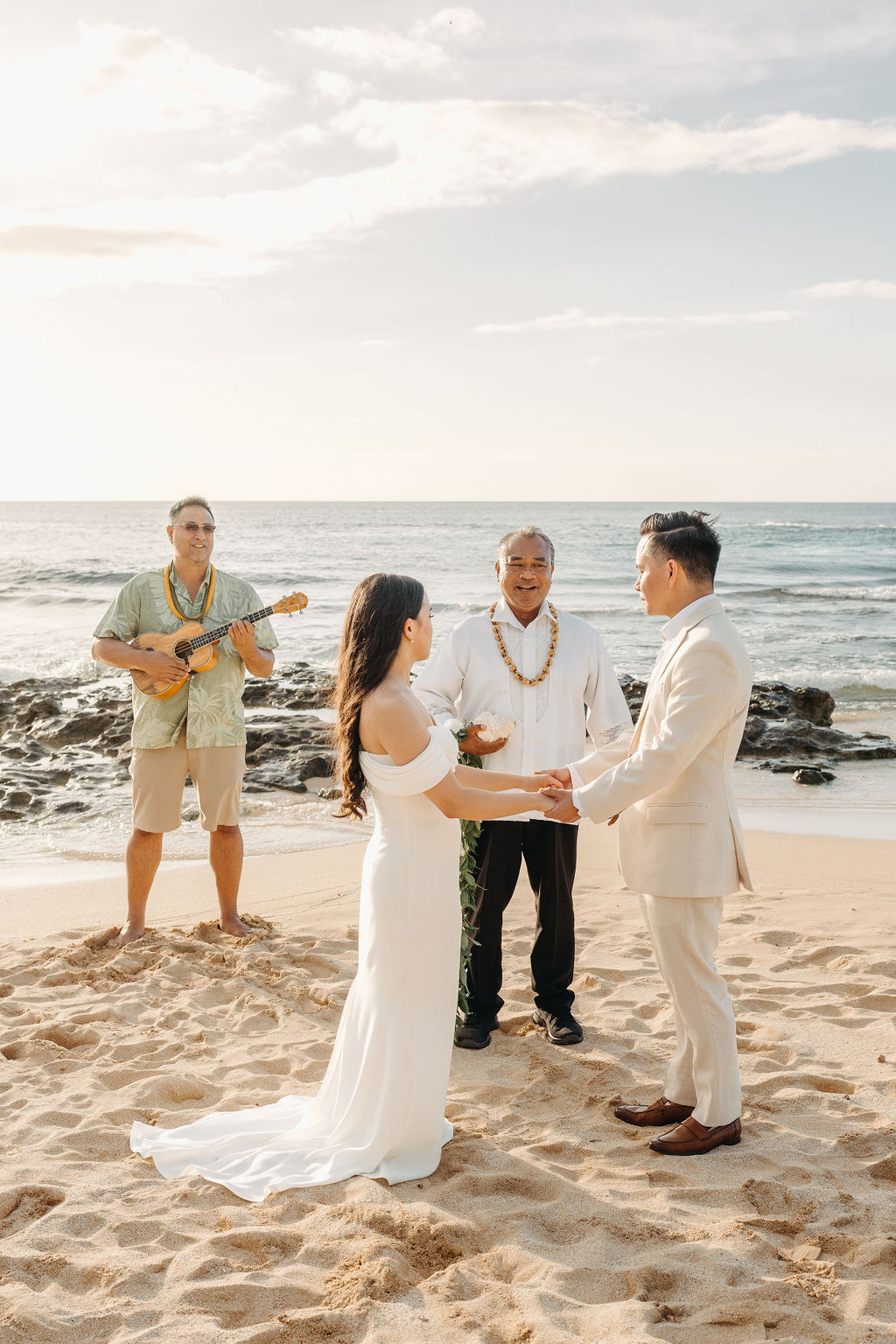 A couple stands holding hands during a beach wedding ceremony, with an officiant and a guitarist in the background by the ocean.