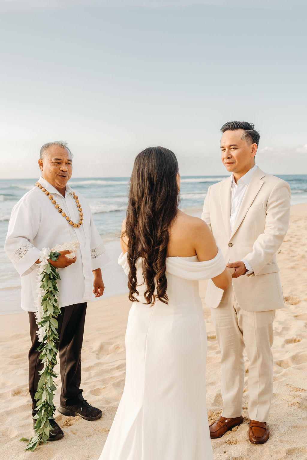 A couple stands holding hands during a beach wedding ceremony, with an officiant and a guitarist in the background by the ocean.