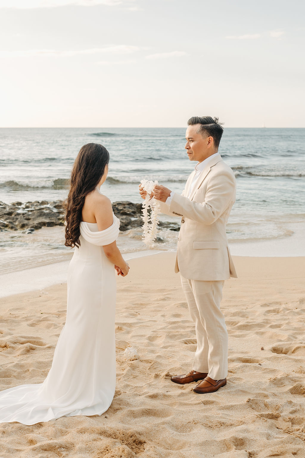 A couple in wedding attire stands on a beach. The man, wearing a light suit, holds a lei while the woman in a white dress faces him. The ocean and rocks are visible in the background.