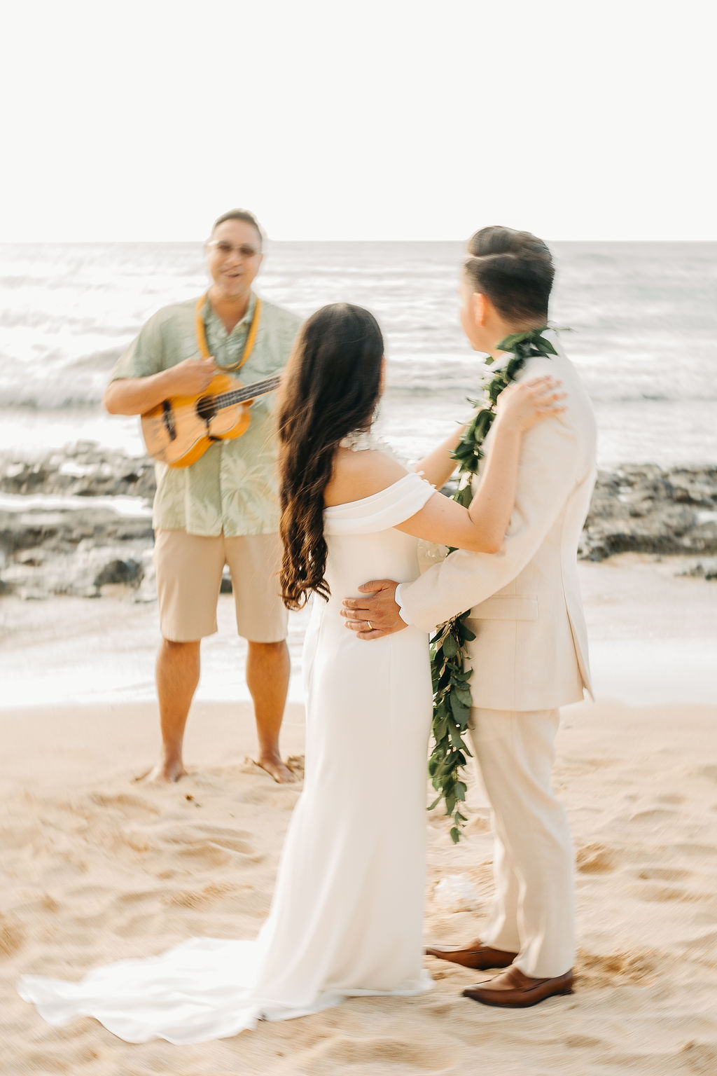  A couple in wedding attire stands on a beach facing an officiant playing a guitar, with the ocean in the background.