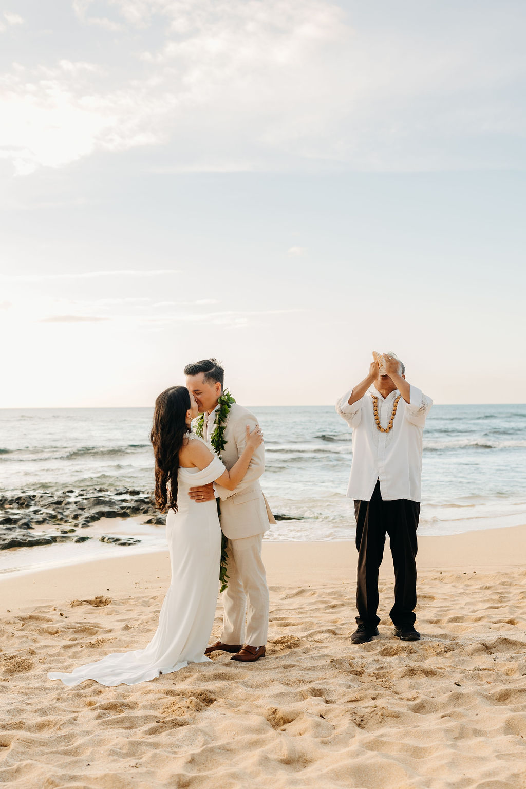 A couple in wedding attire stands on a beach facing an officiant playing a guitar, with the ocean in the background.