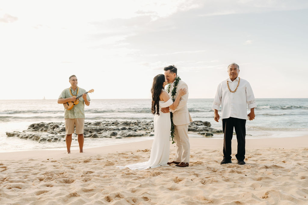 A couple in wedding attire stands on a beach facing an officiant playing a guitar, with the ocean in the background.