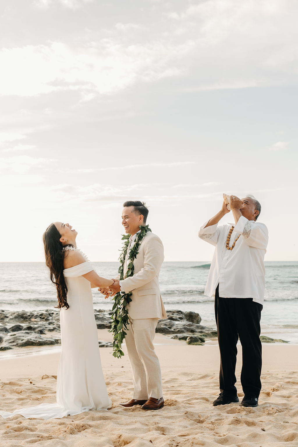 A couple in wedding attire stands on a beach facing an officiant playing a guitar, with the ocean in the background.