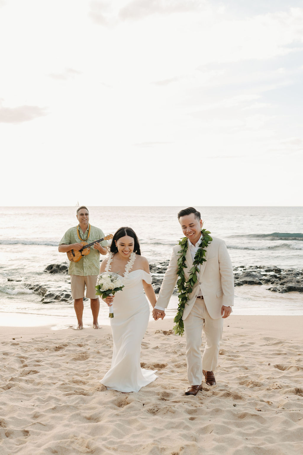 A couple in wedding attire stands on a beach facing an officiant playing a guitar, with the ocean in the background.