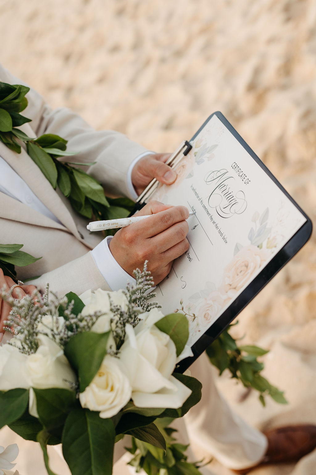 Person in a light suit signs a document on a clipboard while holding a bouquet of white flowers on a sandy beach.