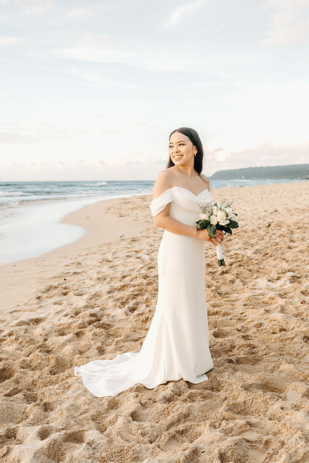 A bride in a white dress stands on a sandy beach, holding a bouquet, with the ocean and sky in the background.