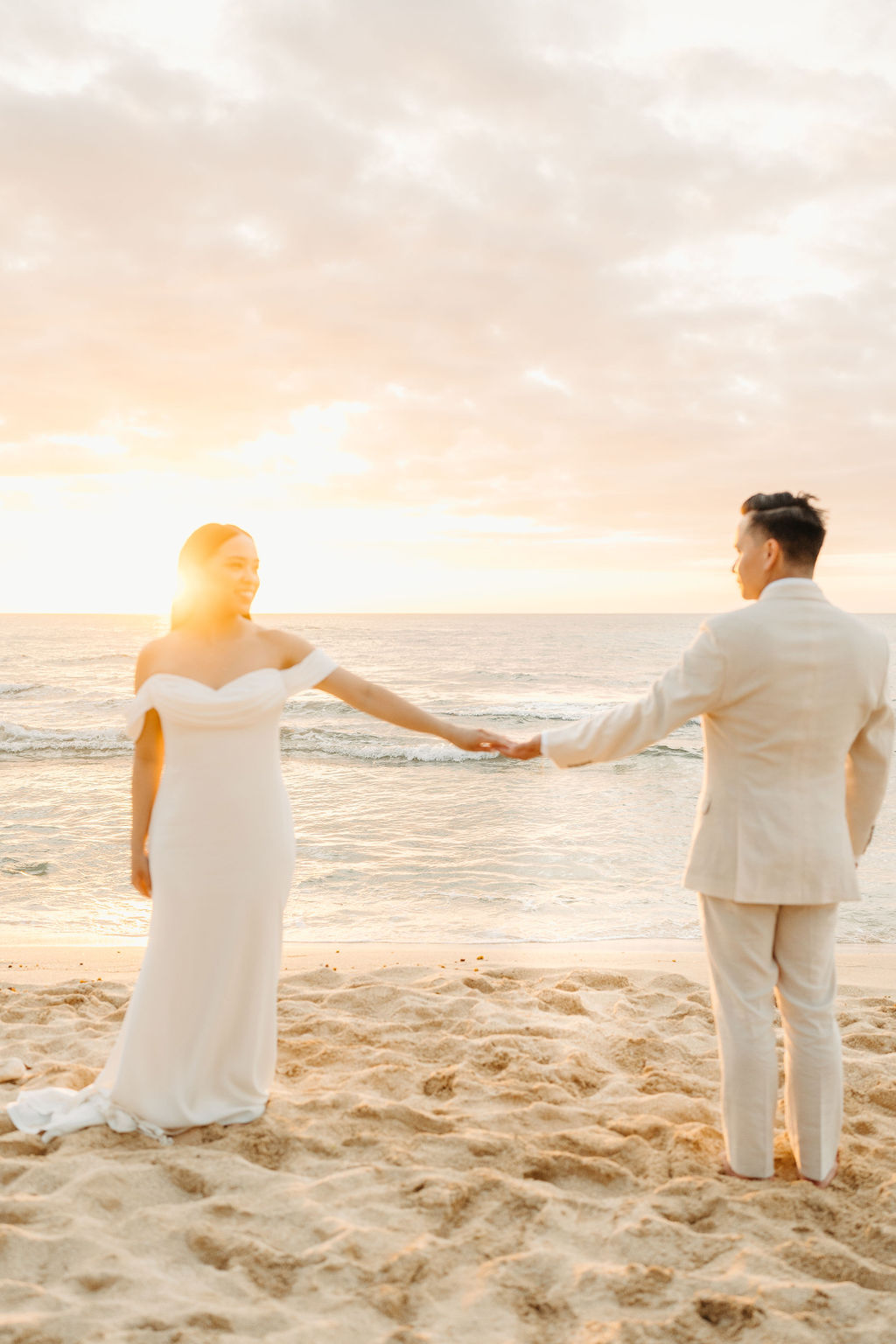 A couple in white attire walks hand in hand on a sandy beach toward the ocean during sunset.