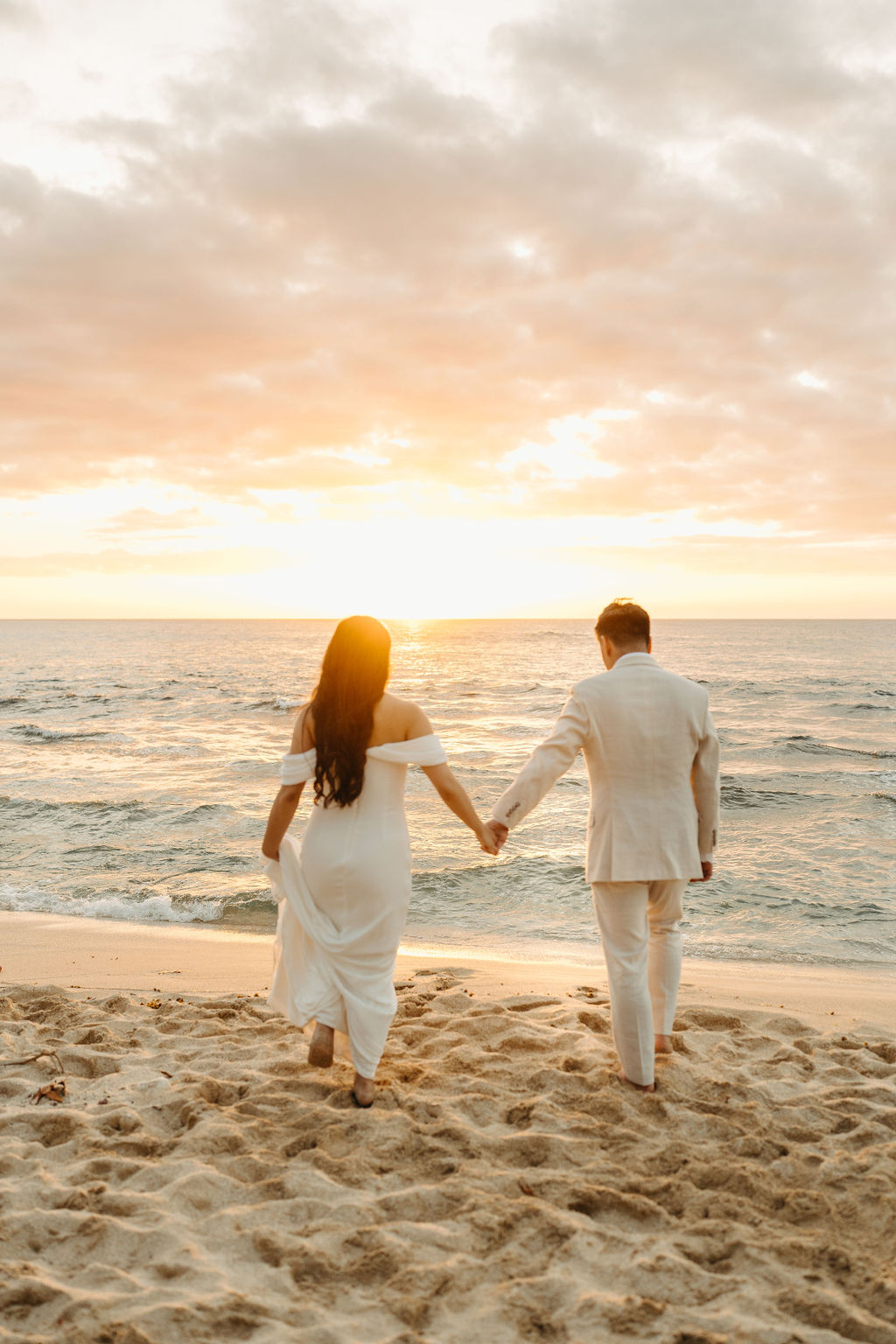 A couple in white attire walks hand in hand on a sandy beach toward the ocean during sunset.