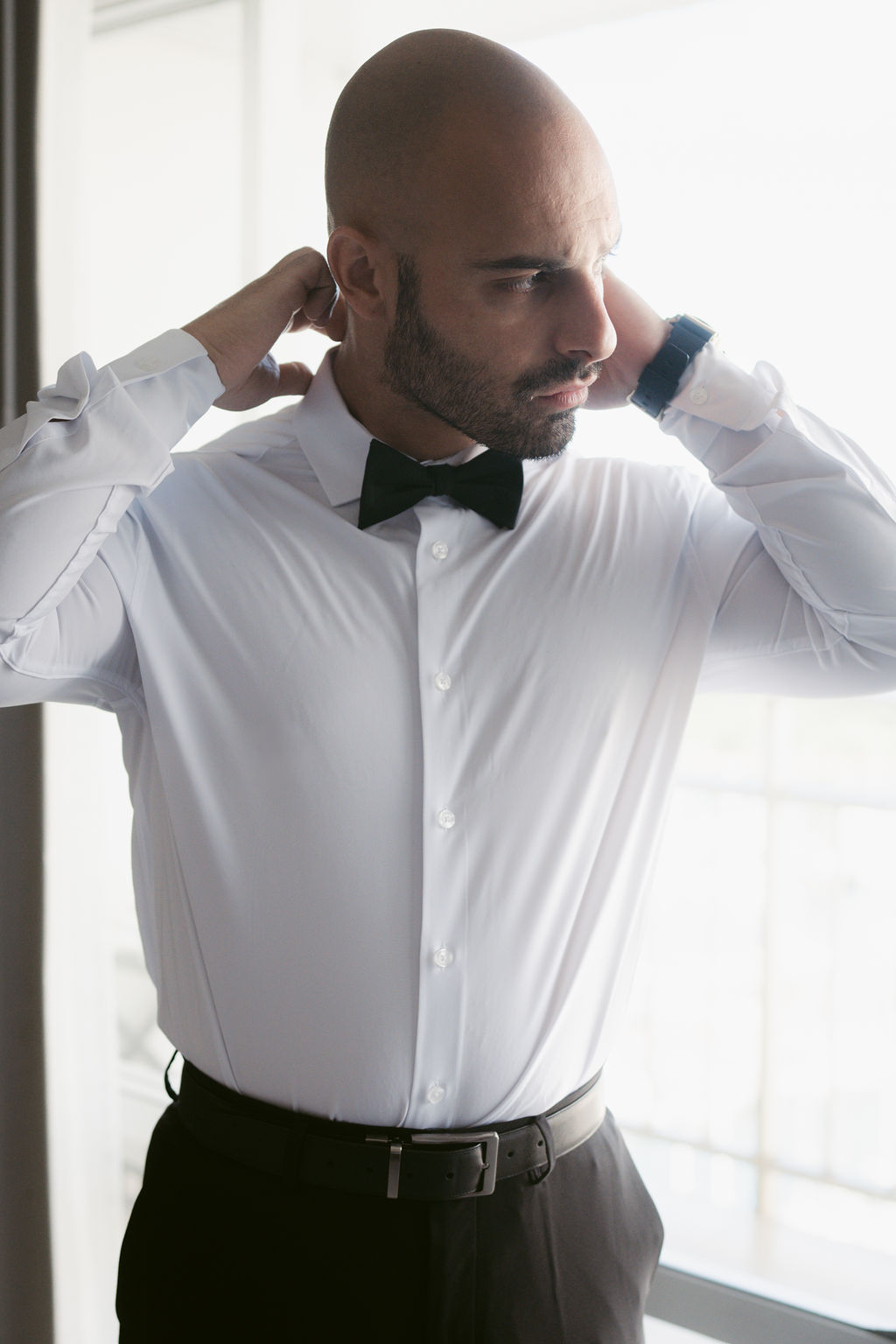 Man in a white dress shirt and black bow tie adjusting his collar, standing by a window.