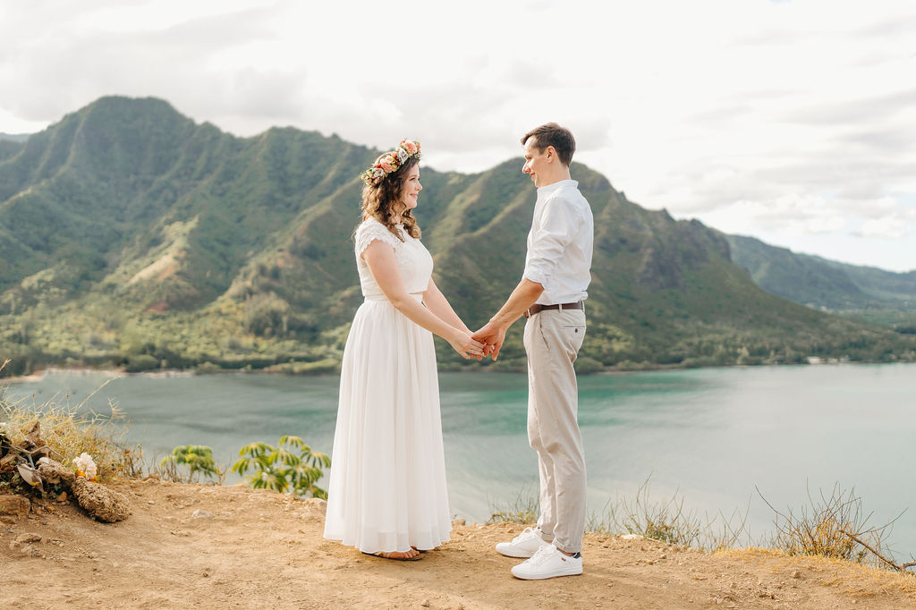 A couple in wedding attire stands holding hands on a cliffside, with mountains and a lake in the background for their hawaii elopement