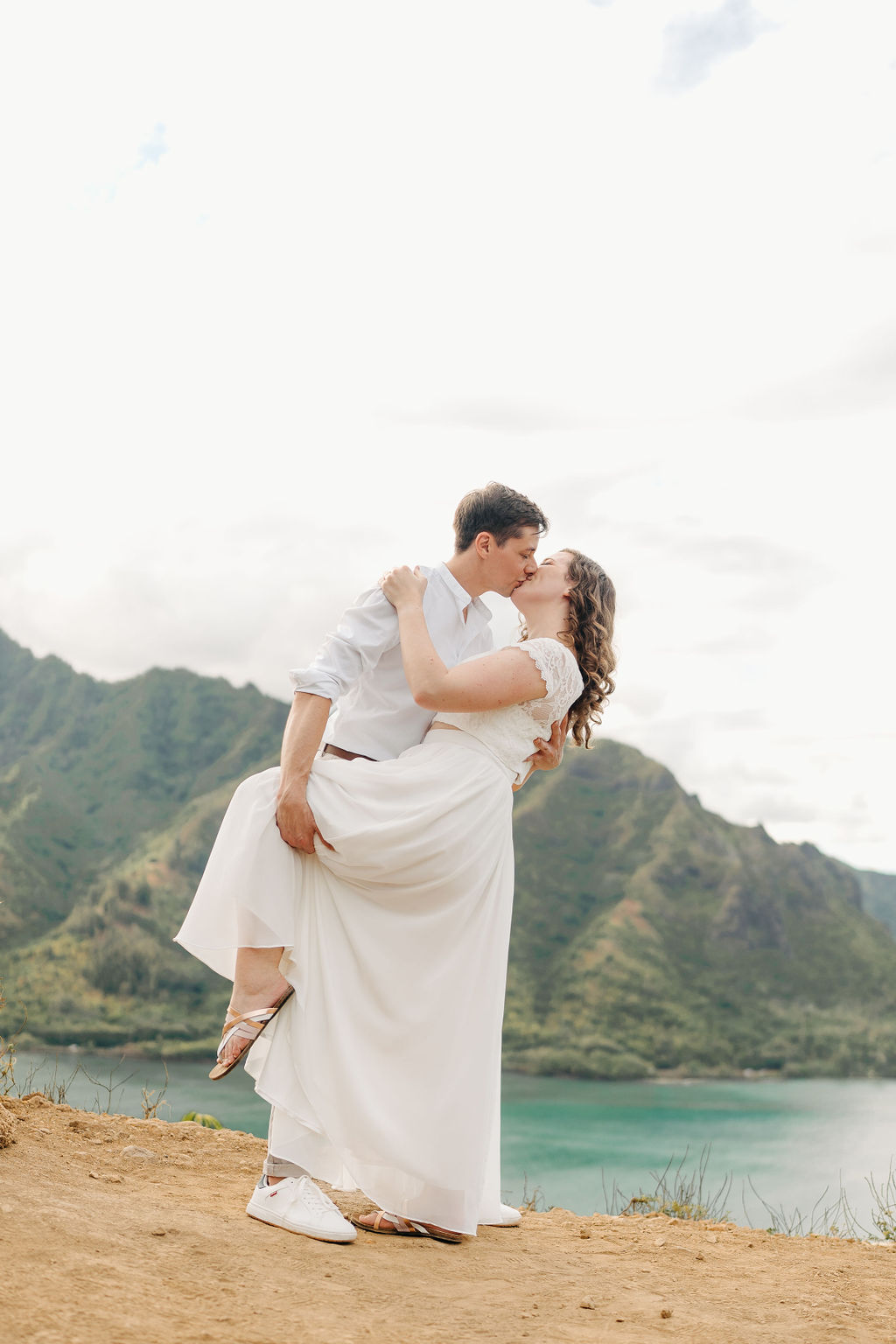 A couple embraces on a cliffside overlooking mountains and a body of water, under a partly cloudy sky for their hawaii elopement 