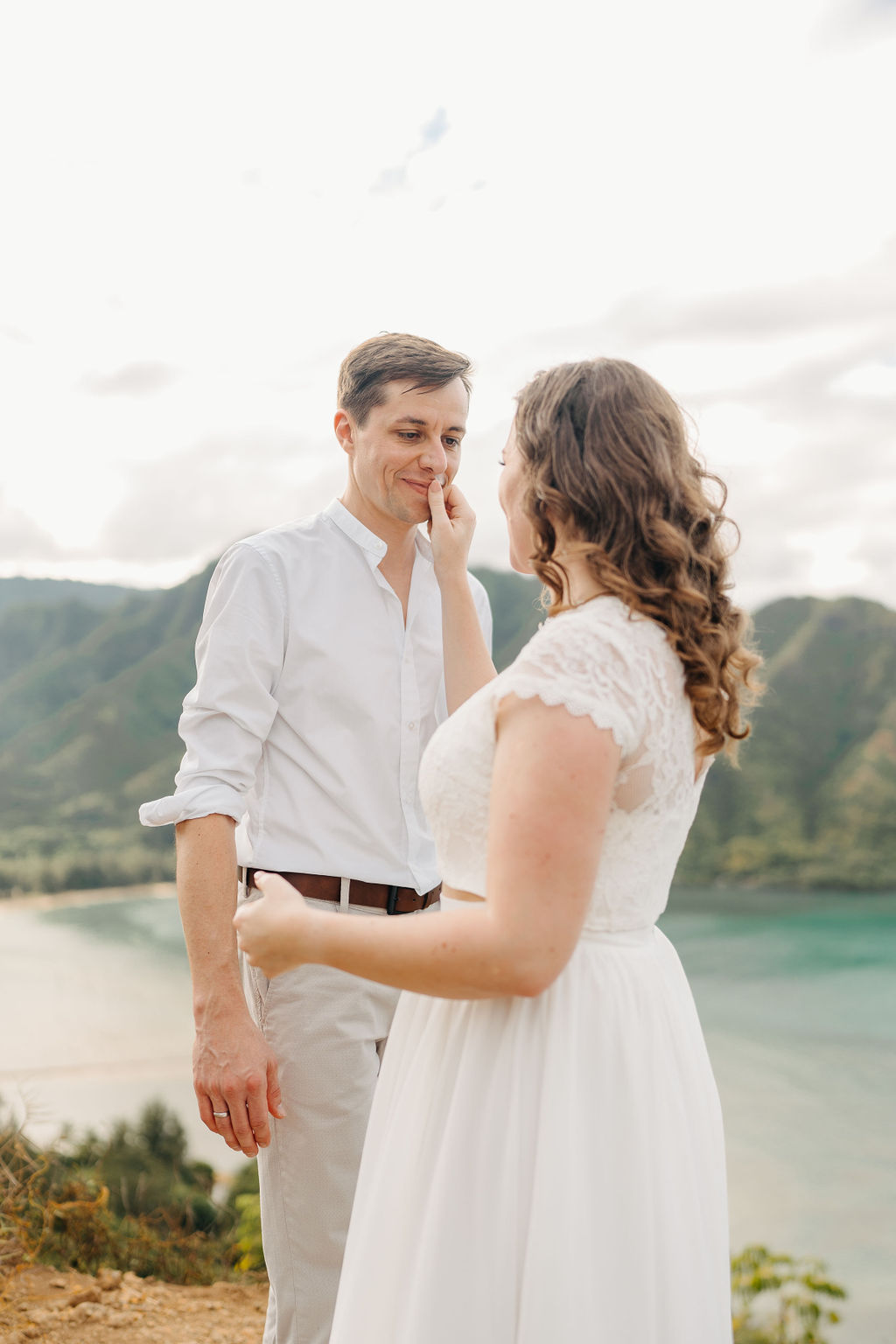 A couple embraces on a cliffside overlooking mountains and a body of water, under a partly cloudy sky for their hawaii elopement 