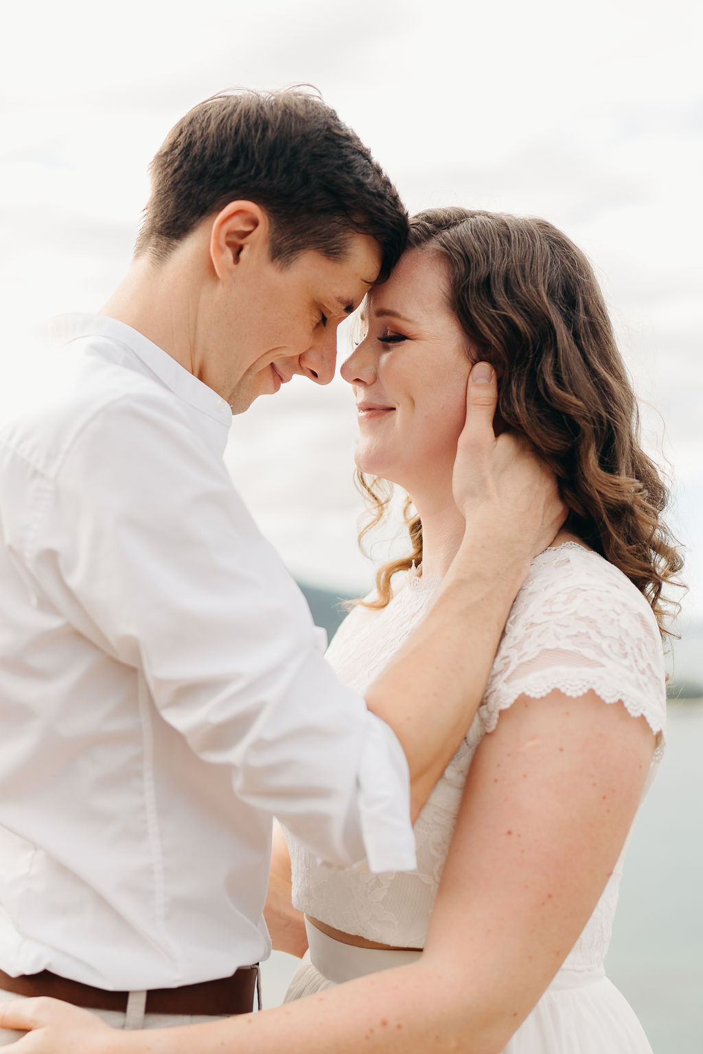 A couple embraces on a cliffside overlooking mountains and a body of water, under a partly cloudy sky for their hawaii elopement 