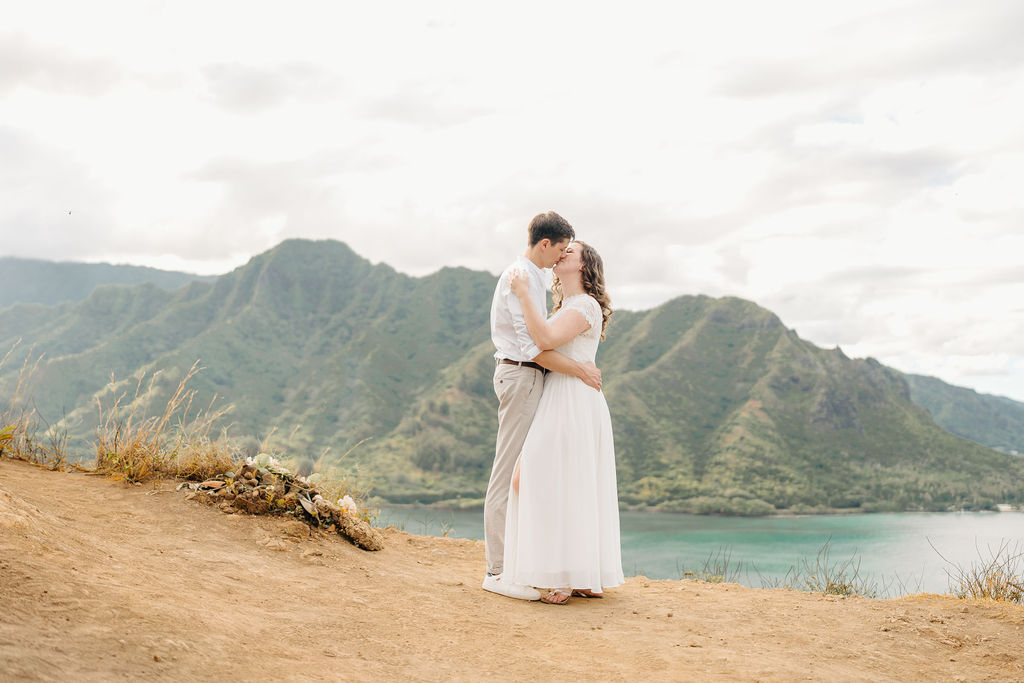 A couple embraces on a cliffside overlooking mountains and a body of water, under a partly cloudy sky for their hawaii elopement 