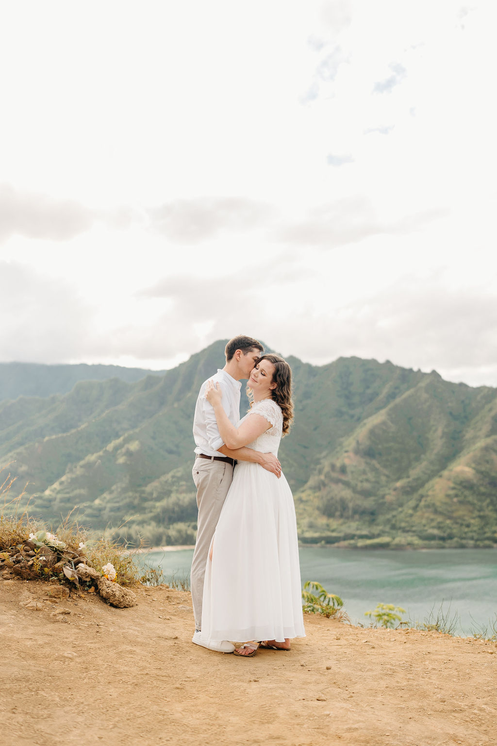 A couple embraces on a cliffside overlooking mountains and a body of water, under a partly cloudy sky for their hawaii elopement 