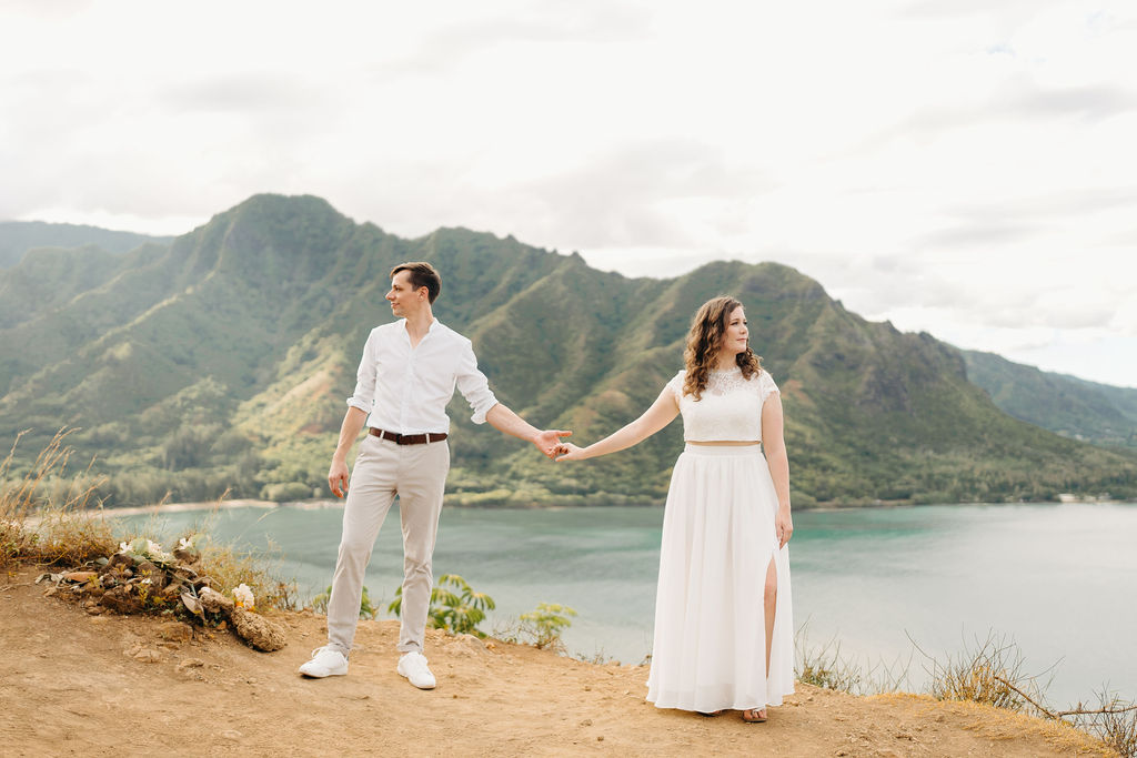 A couple embraces on a cliffside overlooking mountains and a body of water, under a partly cloudy sky