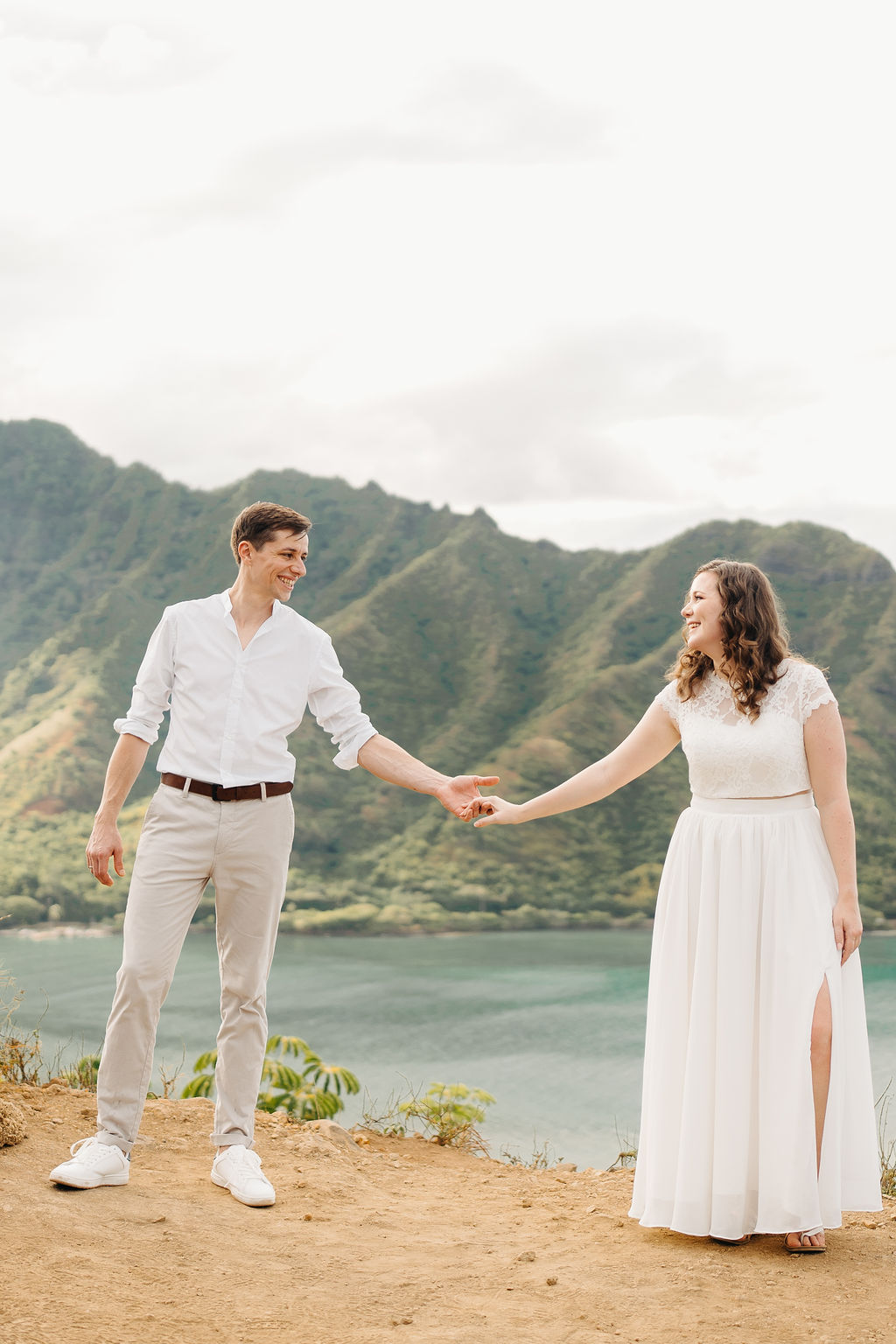 A couple embraces on a cliffside overlooking mountains and a body of water, under a partly cloudy sky for their hawaii elopement 