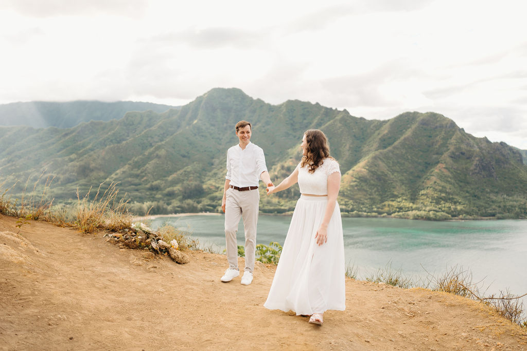 A couple embraces on a cliffside overlooking mountains and a body of water, under a partly cloudy sky for their hawaii elopement