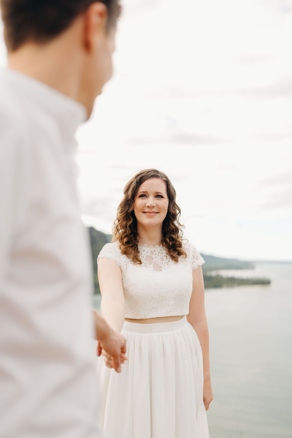 A couple embraces on a cliffside overlooking mountains and a body of water, under a partly cloudy sky for their hawaii elopement