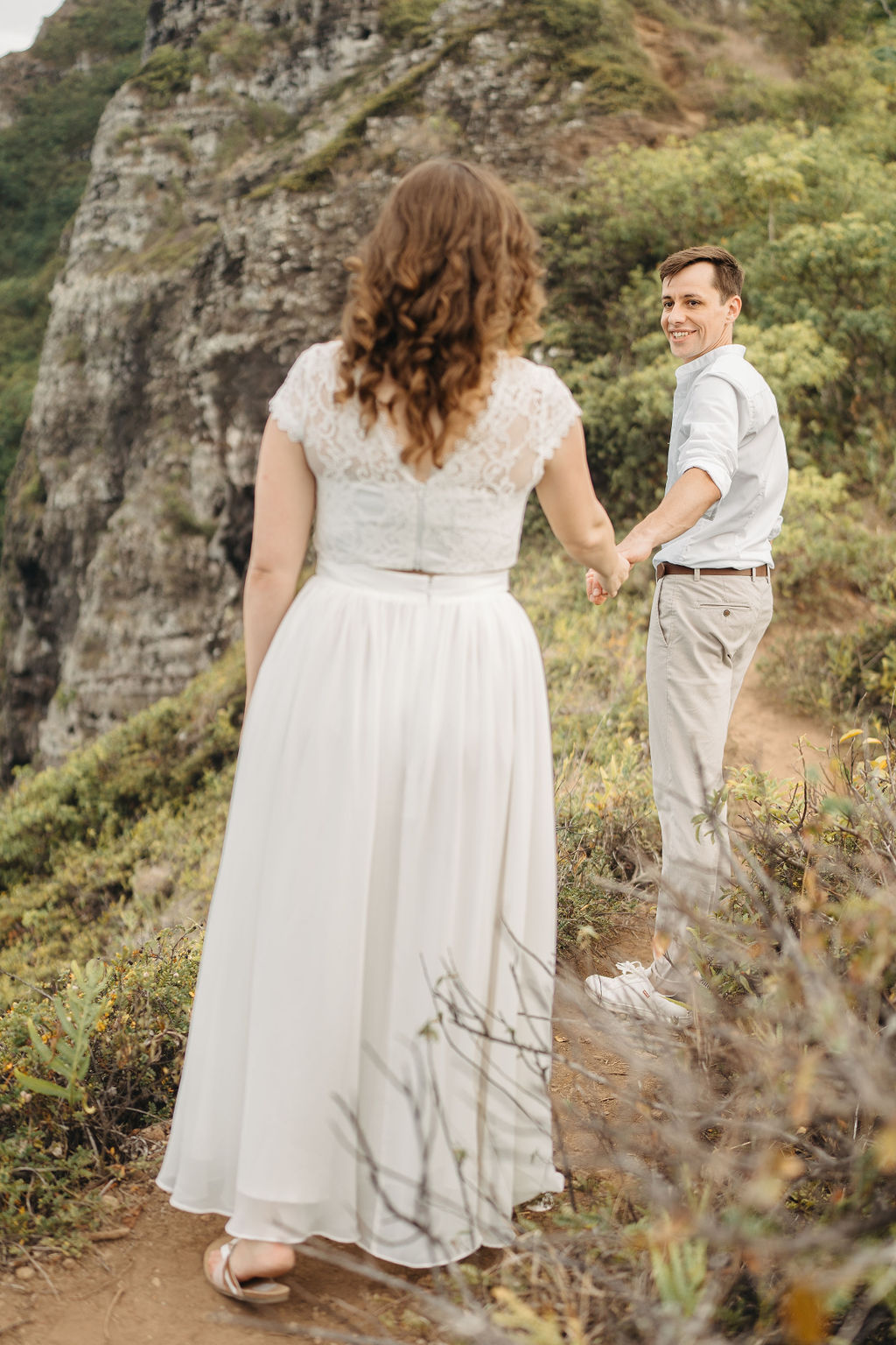 A couple embraces on a cliffside overlooking mountains and a body of water, under a partly cloudy sky for their hawaii elopement