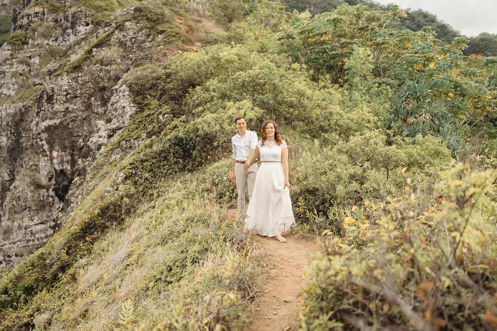 A couple embraces on a cliffside overlooking mountains and a body of water, under a partly cloudy sky