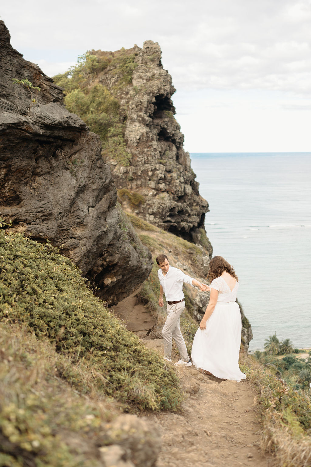 A couple embraces on a cliffside overlooking mountains and a body of water, under a partly cloudy sky for their hawaii elopement