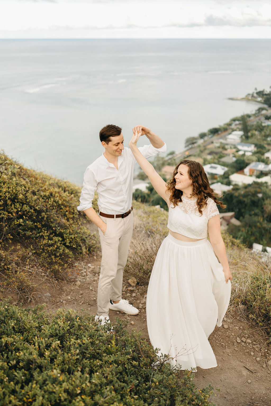 A couple embraces on a cliffside overlooking mountains and a body of water, under a partly cloudy sky for their hawaii elopement