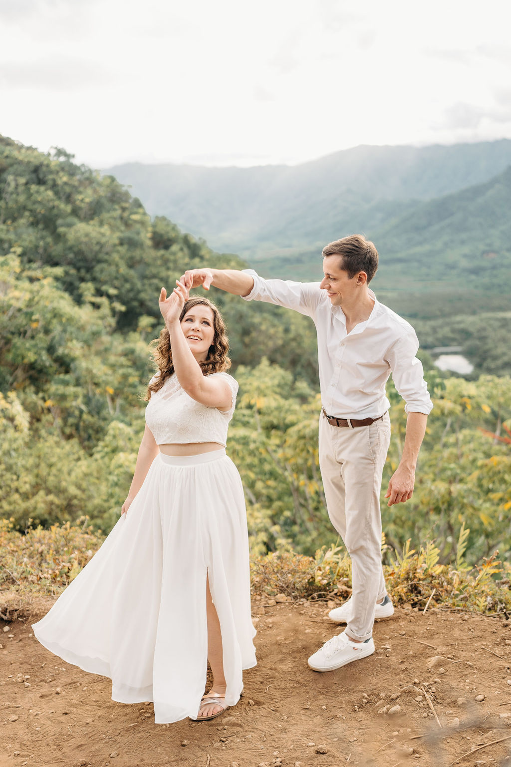 A couple embraces on a cliffside overlooking mountains and a body of water, under a partly cloudy sky