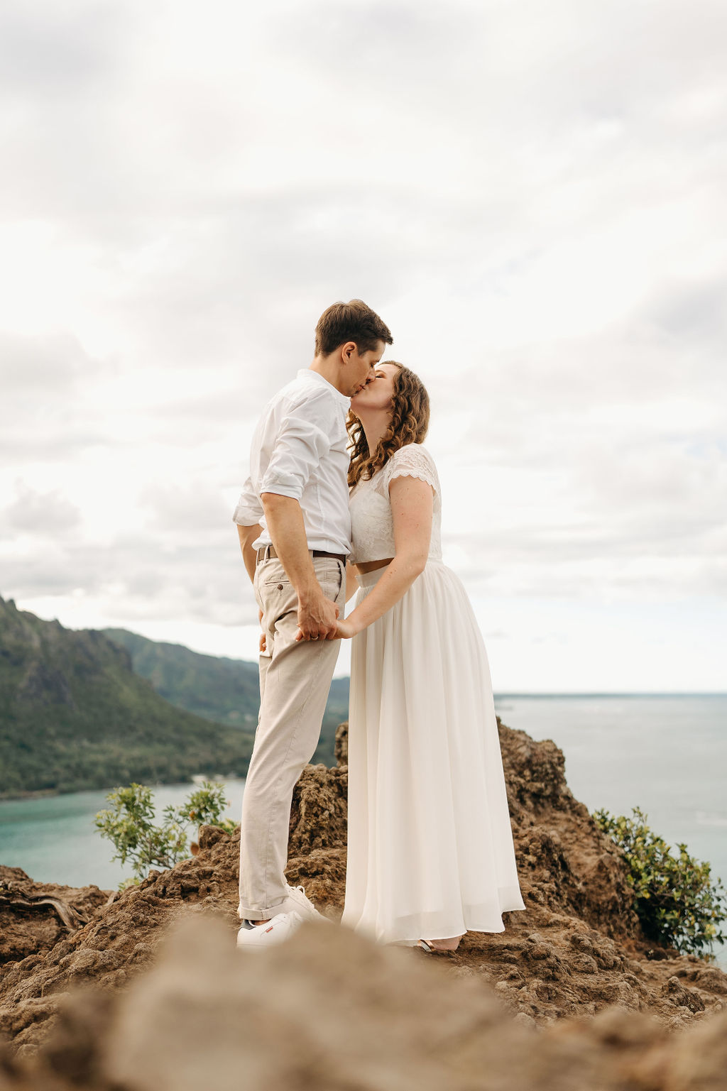 A couple embraces on a cliffside overlooking mountains and a body of water, under a partly cloudy sky 