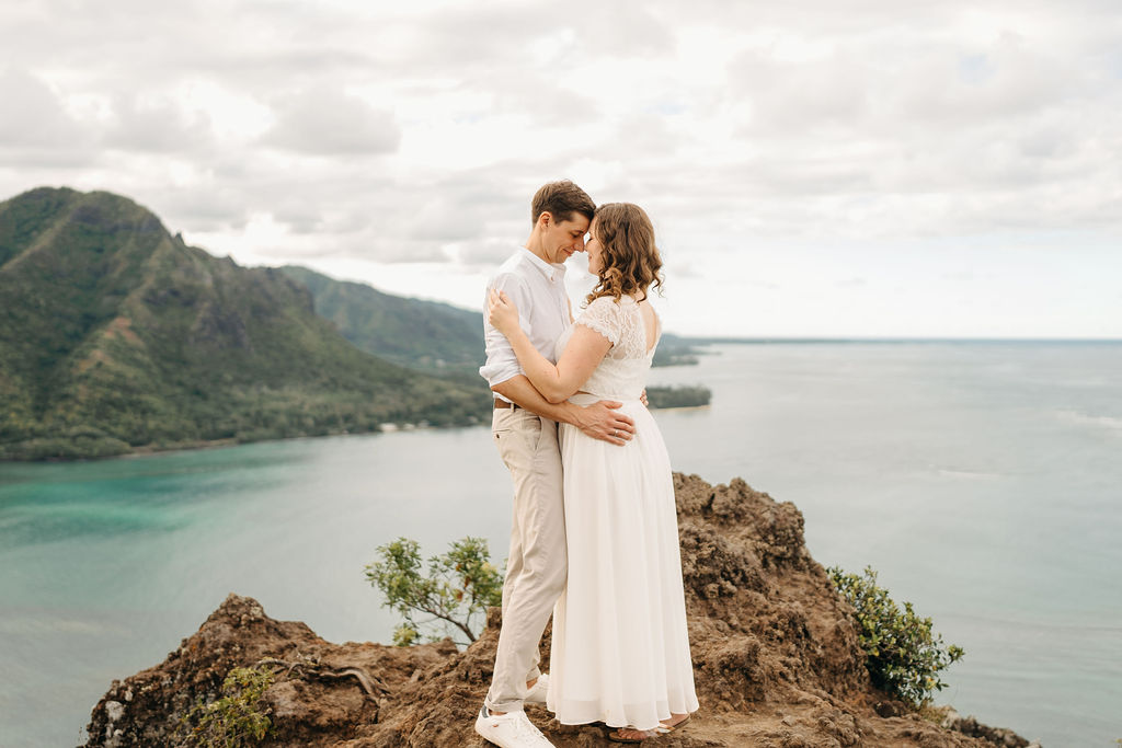 A couple embraces on a cliffside overlooking mountains and a body of water, under a partly cloudy sky 