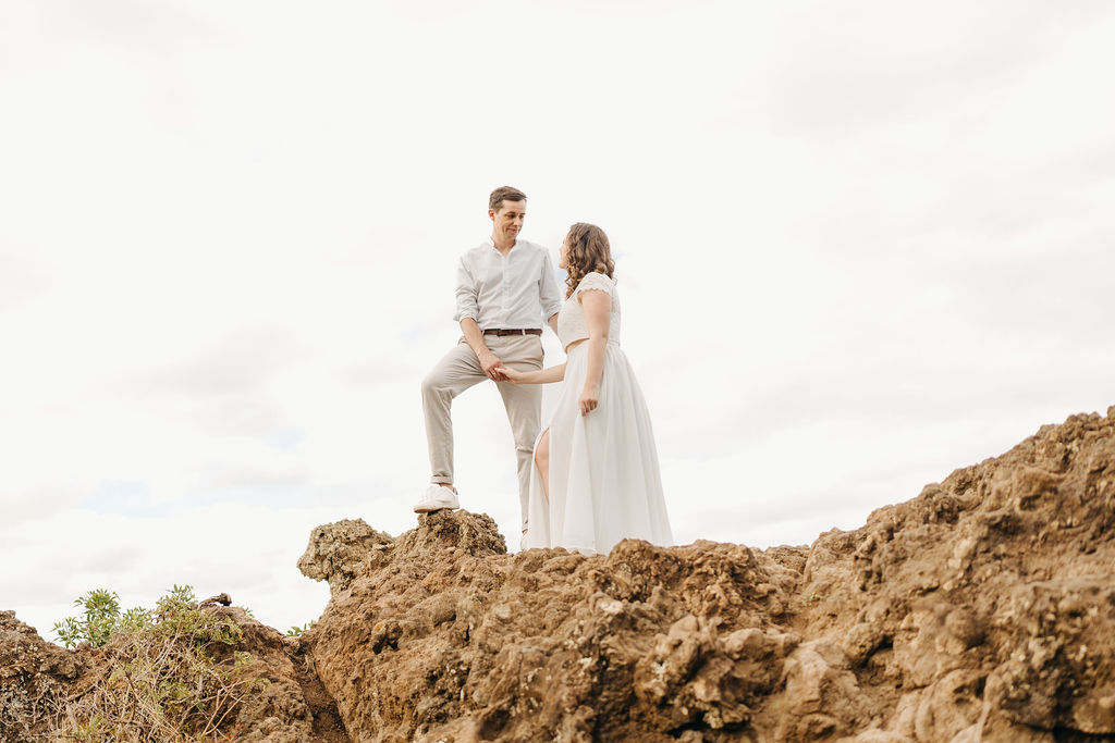 A couple embraces on a cliffside overlooking mountains and a body of water, under a partly cloudy sky for their hawaii elopement