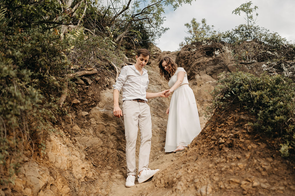 A couple in light clothing descends a rocky slope, holding hands. Dense greenery surrounds them under a cloudy sky.