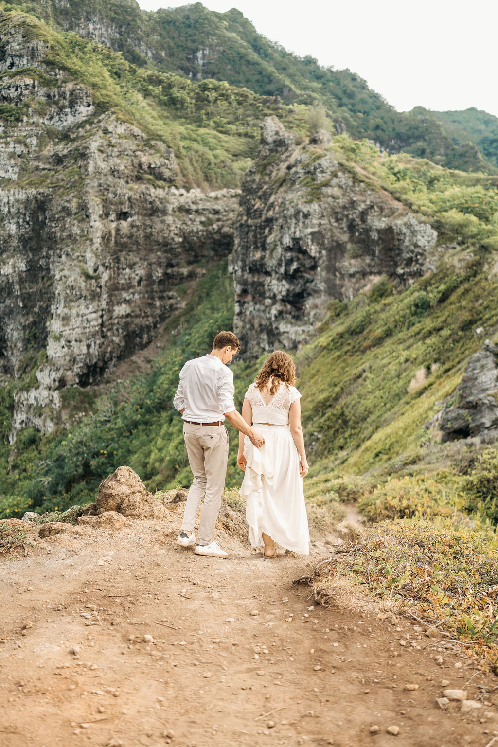 A couple embraces on a cliffside overlooking mountains and a body of water, under a partly cloudy sky 