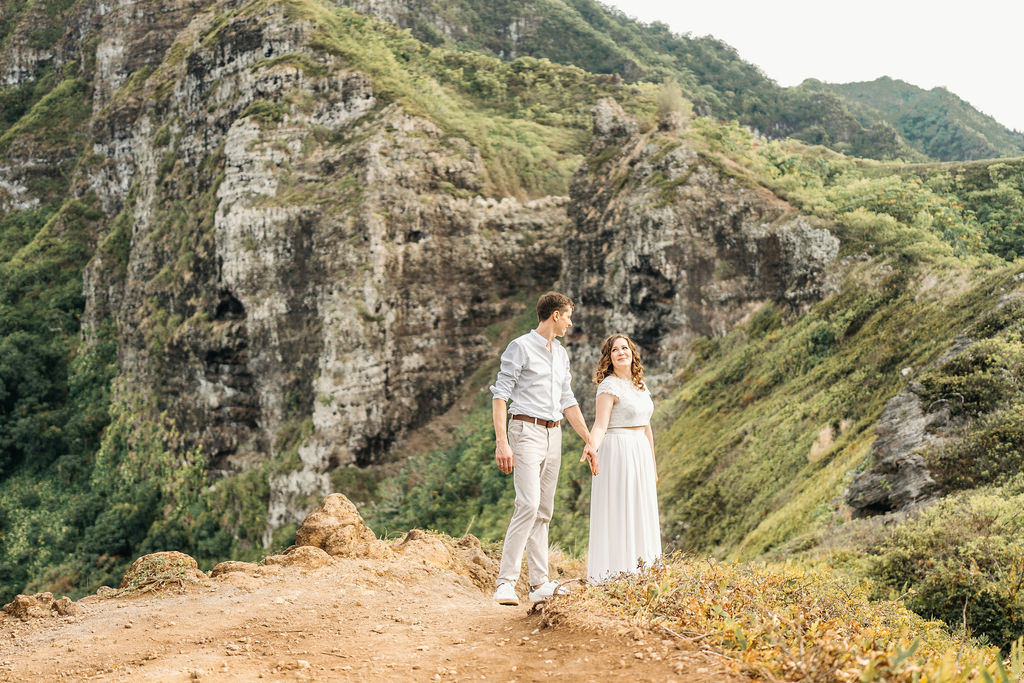 A couple in light clothing descends a rocky slope, holding hands. Dense greenery surrounds them under a cloudy sky.