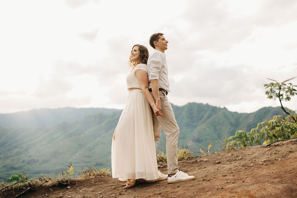 A couple embraces on a cliffside overlooking mountains and a body of water, under a partly cloudy sky