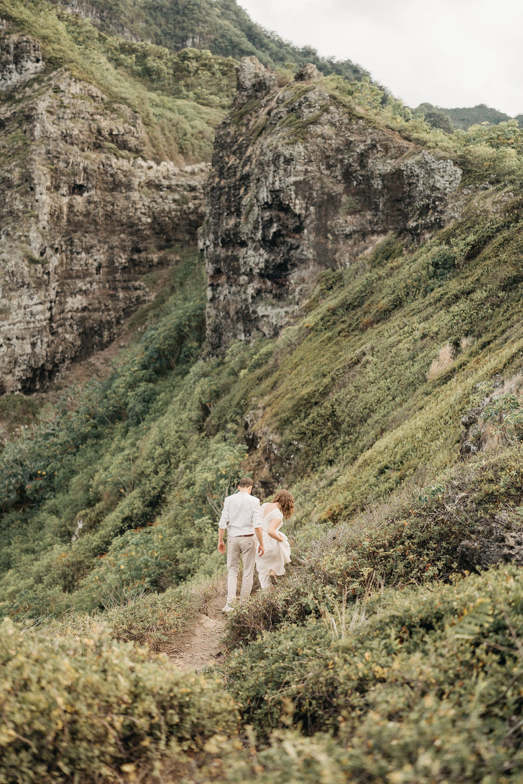 A couple embraces on a cliffside overlooking mountains and a body of water, under a partly cloudy sky 