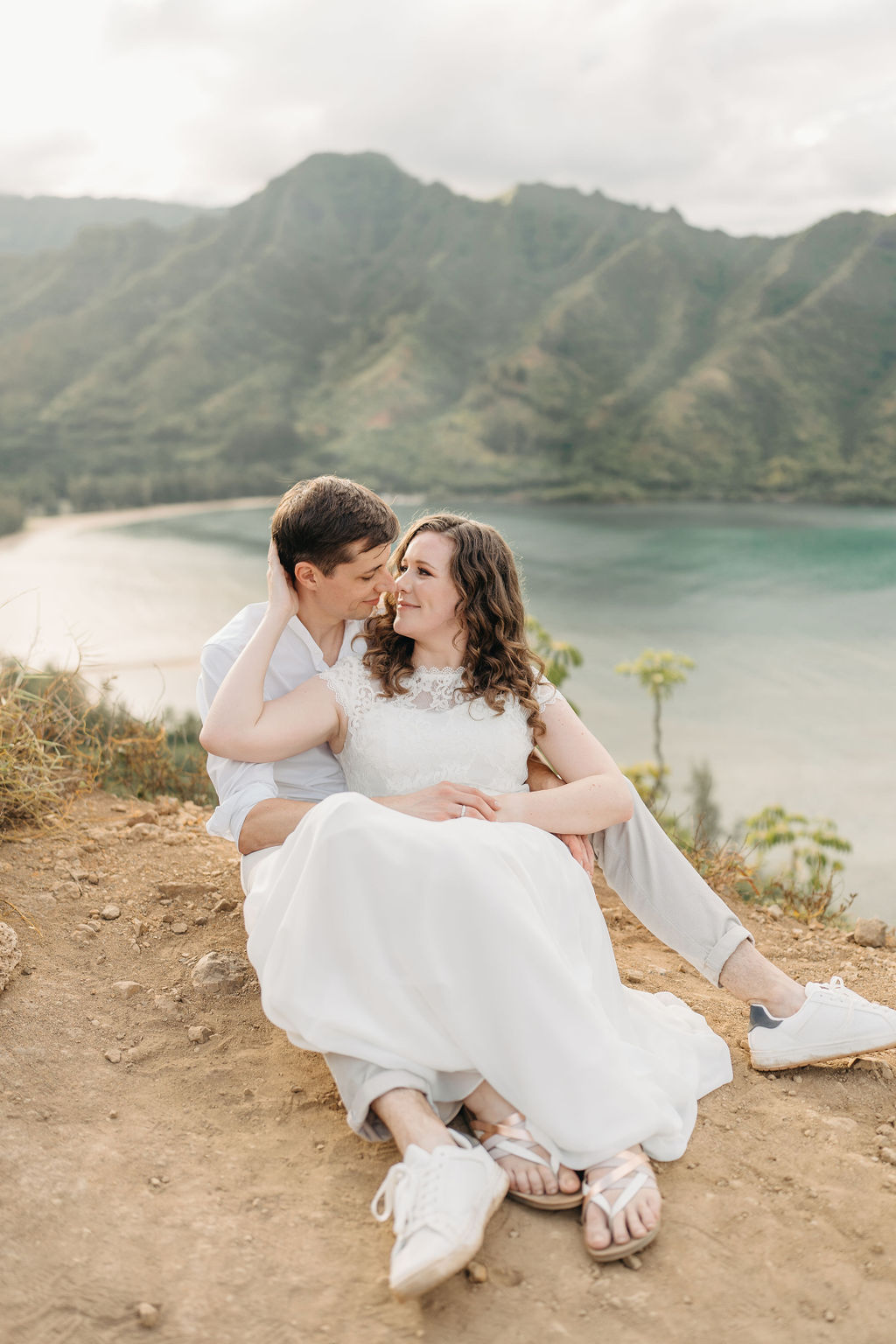 A couple embraces on a cliffside overlooking mountains and a body of water, under a partly cloudy sky