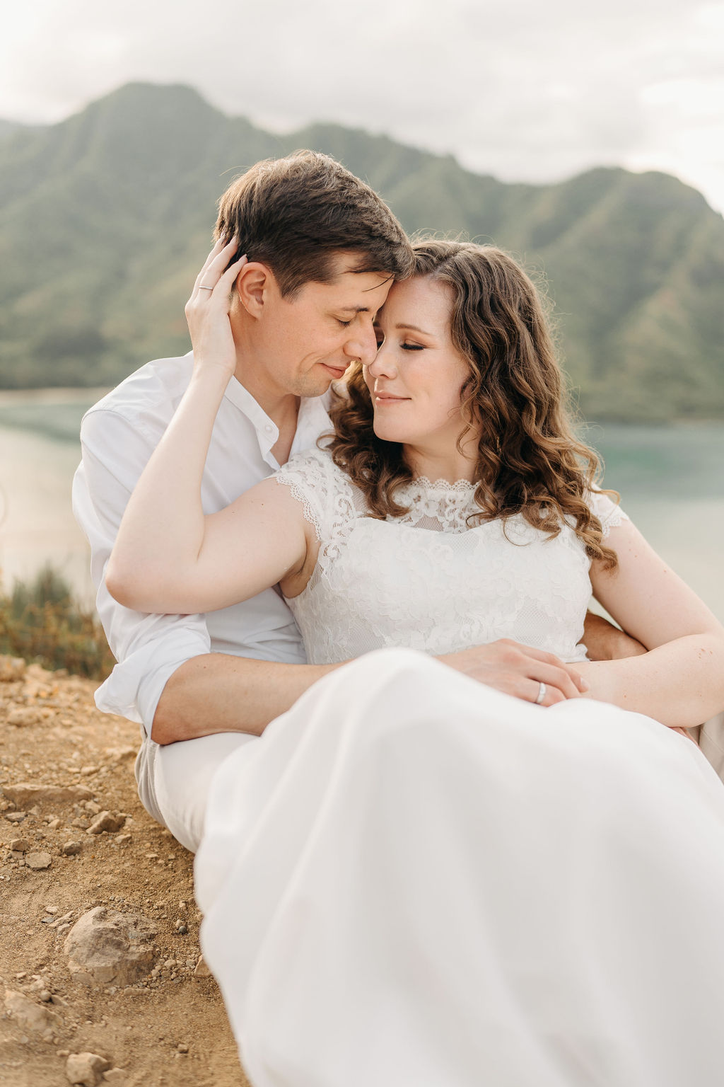 A couple embraces on a cliffside overlooking mountains and a body of water, under a partly cloudy sky