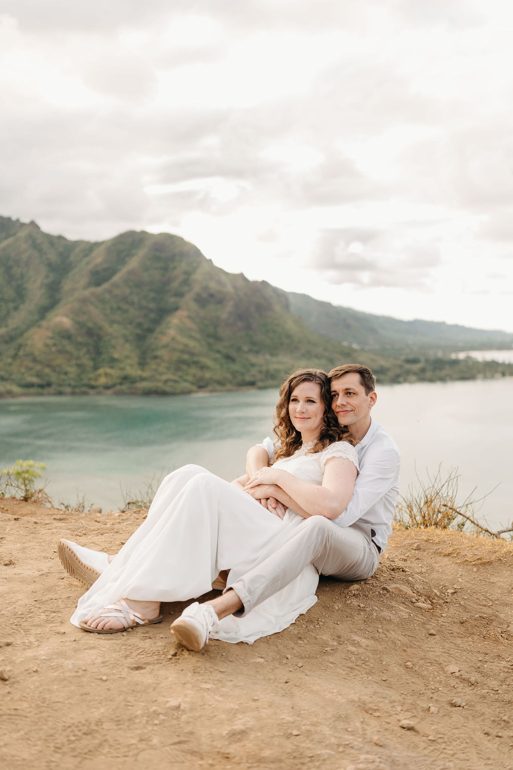 A couple embraces on a cliffside overlooking mountains and a body of water, under a partly cloudy sky