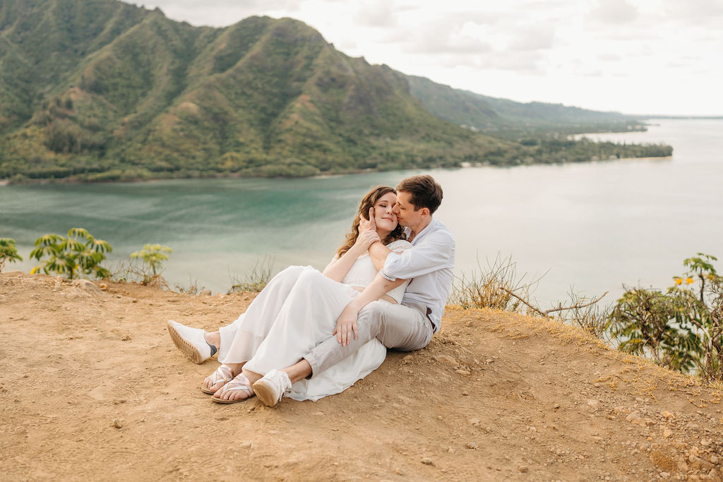 A couple embraces on a cliffside overlooking mountains and a body of water, under a partly cloudy sky 