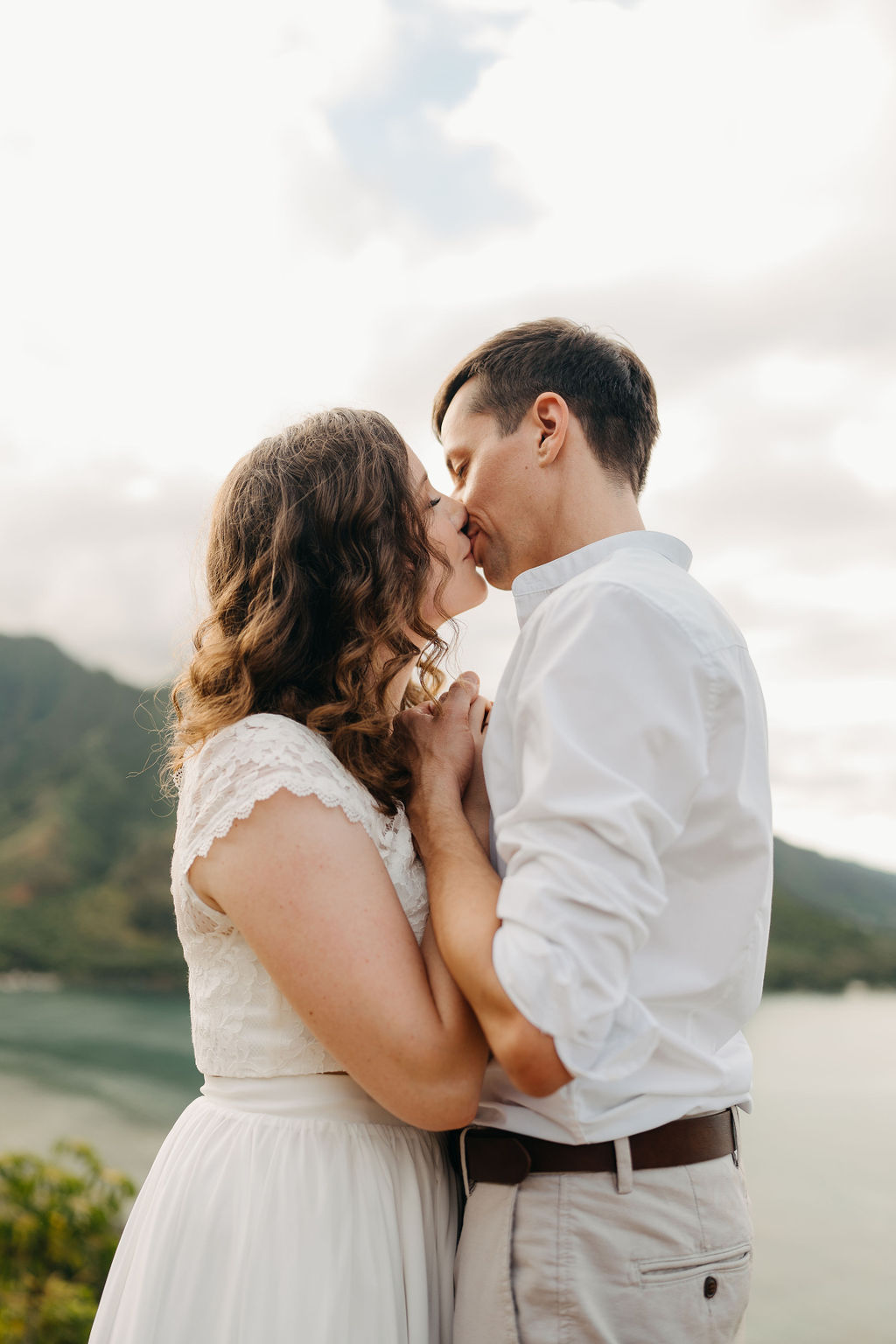 A couple embraces on a cliffside overlooking mountains and a body of water, under a partly cloudy sky