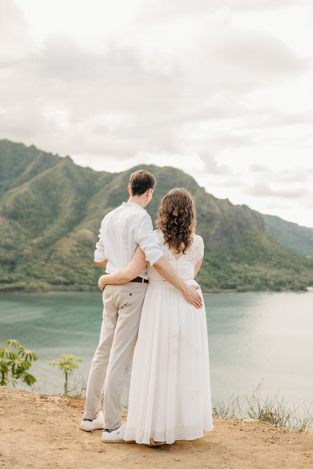 A couple embraces on a cliffside overlooking mountains and a body of water, under a partly cloudy sky