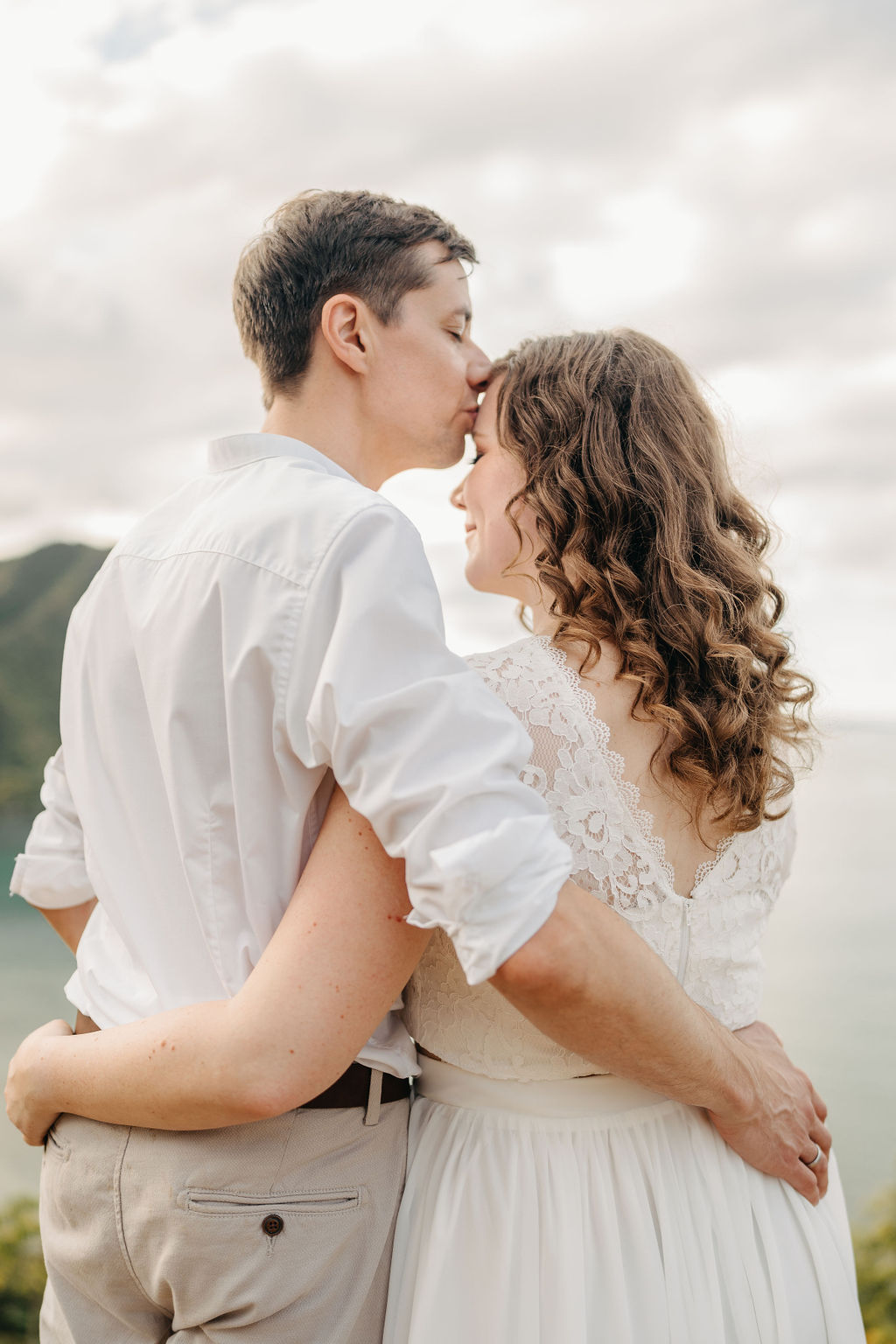 A couple embraces on a cliffside overlooking mountains and a body of water, under a partly cloudy sky