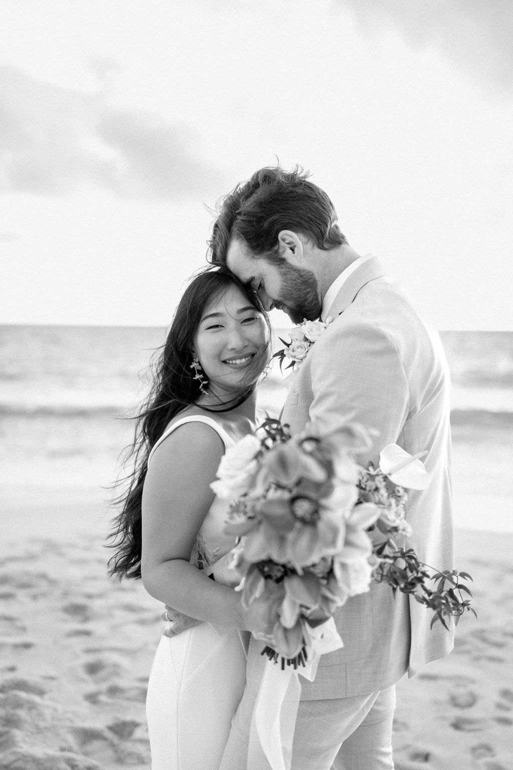 A couple in wedding attire embraces on a beach, with the ocean and sky in the background. The woman holds a bouquet and both appear happy for their Waimanalo Beach elopement 